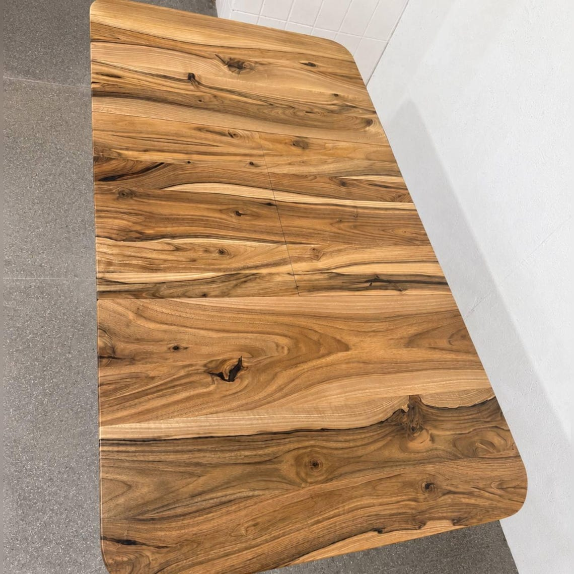 A rectangular wooden tabletop with rounded corners, showcasing a rich natural wood grain pattern in warm brown and golden tones, photographed in a clean indoor setting.