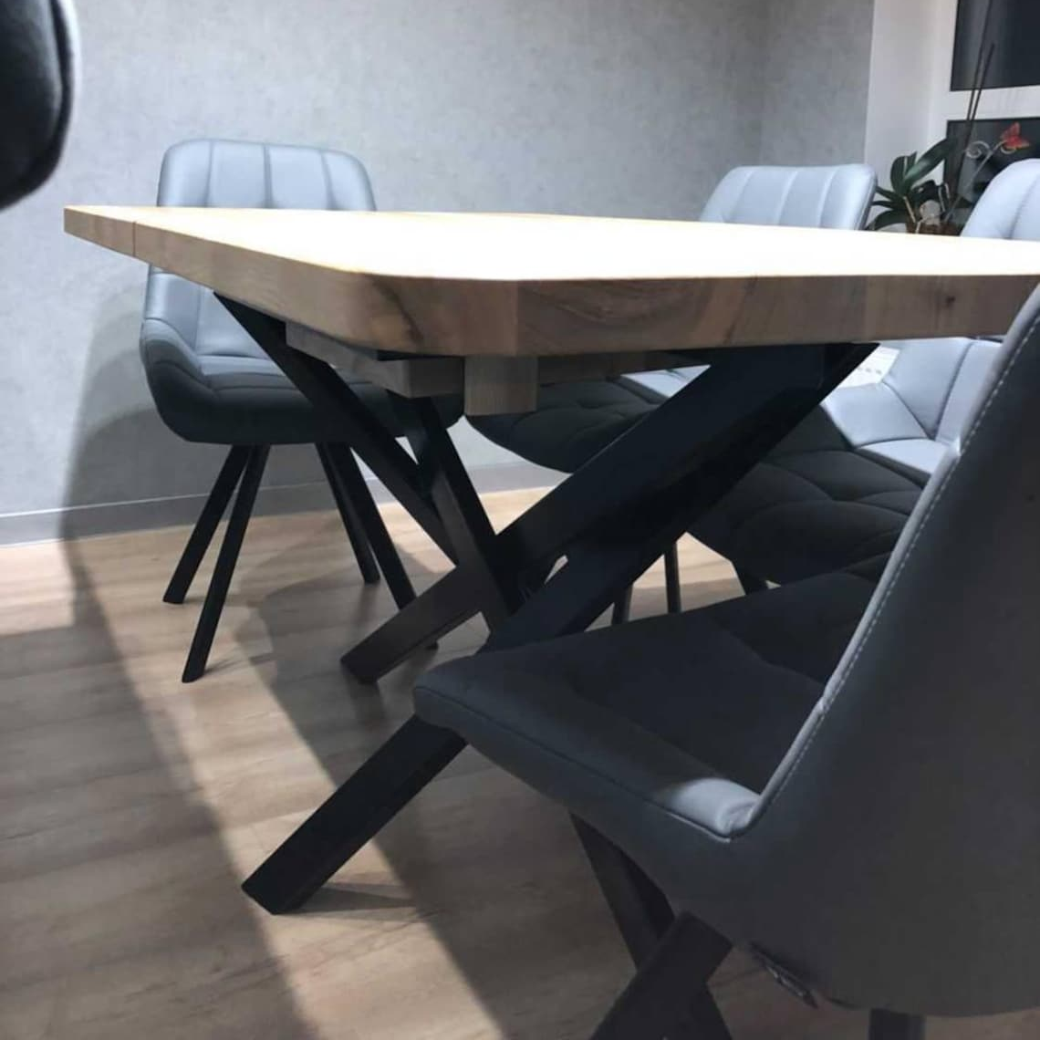 A modern dining table with a light natural wood top and black metal X-shaped legs, surrounded by gray upholstered chairs in a minimalist room with a textured gray wall and light wood floor.