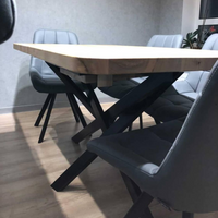 A modern dining table with a light natural wood top and black metal X-shaped legs, surrounded by gray upholstered chairs in a minimalist room with a textured gray wall and light wood floor.