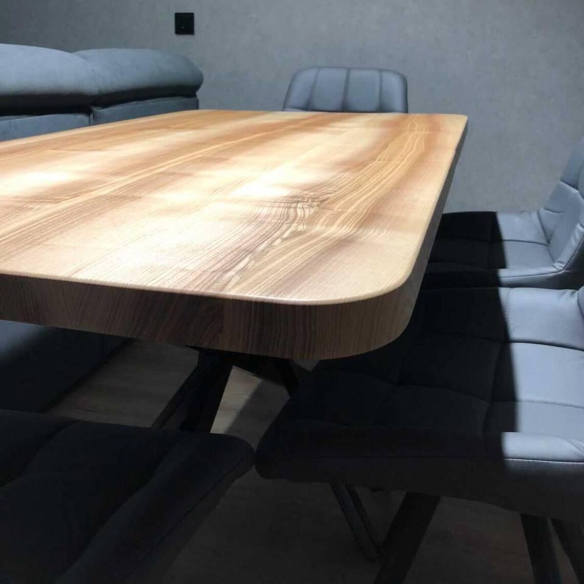 A modern wooden dining table with rounded corners and a light natural wood grain finish, surrounded by black and light gray upholstered chairs in a minimalist room with a gray wall.