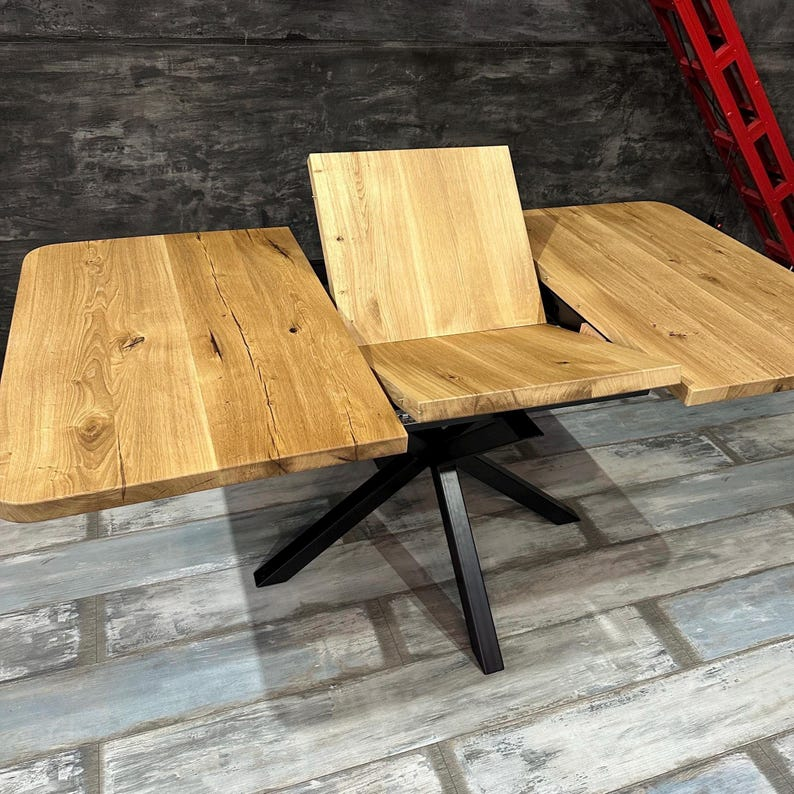 A modern wooden dining table with a black metal star base, shown partially unfolded to reveal its leaf design. The tabletops and backrest are made of natural light brown wood with prominent grain patterns.