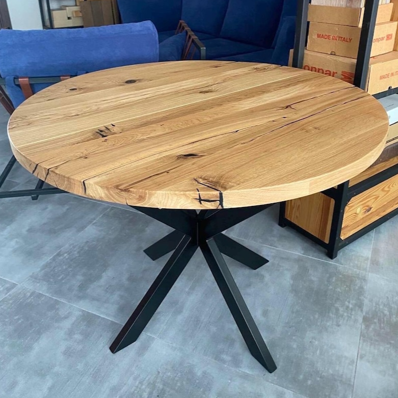 A round wooden dining table with a natural oak finish and black metal cross base, shown in a furniture showroom.