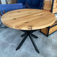 A round wooden dining table with a natural oak finish and black metal cross base, shown in a furniture showroom.
