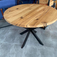 Round wooden dining table with a natural oak top and a black metal star base, shown in a warehouse or showroom setting on a gray tiled floor.