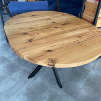 A round wooden dining table with a natural oak finish and black metal legs, shown in a showroom setting on a gray tiled floor.