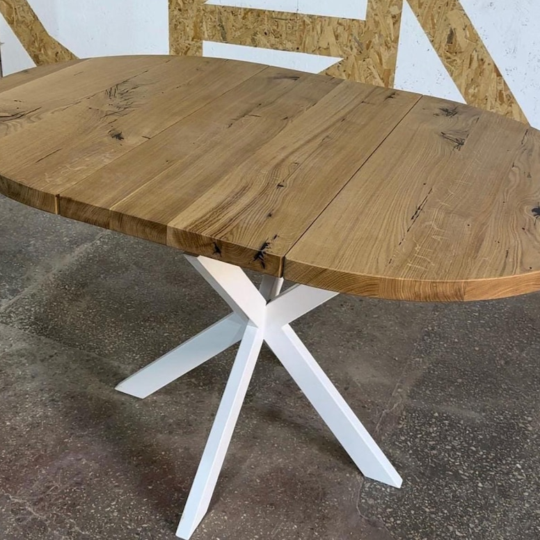 An oval wooden dining table with a natural oak-look top and a white painted X-shaped metal pedestal base, shown in an indoor warehouse or showroom setting.