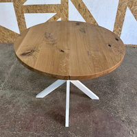 A round wooden dining table with a natural oak finish and a white metal tripod base, photographed in a warehouse setting.