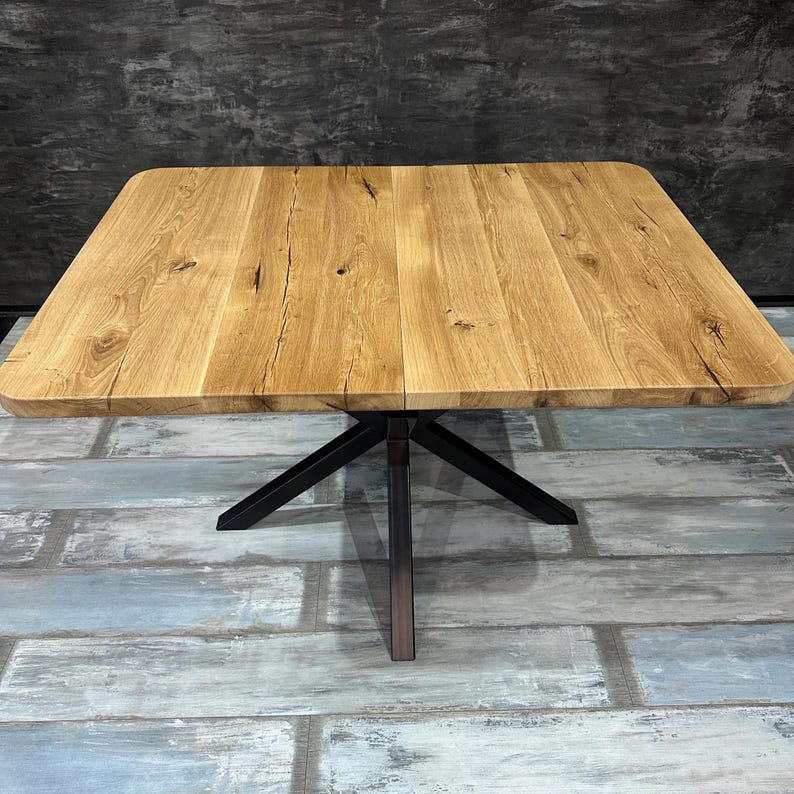 A square wooden dining table with a natural oak top and a black metal X-shaped pedestal base, set against a dark textured wall and distressed gray wood flooring.