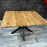 A square wooden dining table with a natural oak-look top and a black metal star base, shown on a rustic gray floor against a dark textured wall.