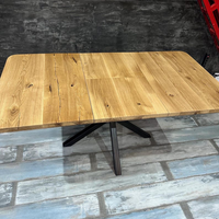 A rectangular wooden dining table with a natural oak finish and a black metal star base, shown in a warehouse or showroom setting with a gray concrete wall and distressed blue-gray floor.
