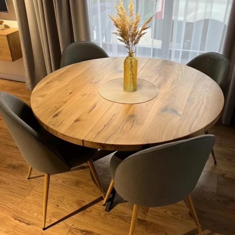 Round wooden dining table with a natural oak finish, surrounded by four gray upholstered chairs with light wood legs, set in a dining room with a glass vase of dried grass on top.