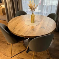 Round wooden dining table with a natural oak finish, surrounded by four gray upholstered chairs with light wood legs, set in a dining room with a glass vase of dried grass on top.
