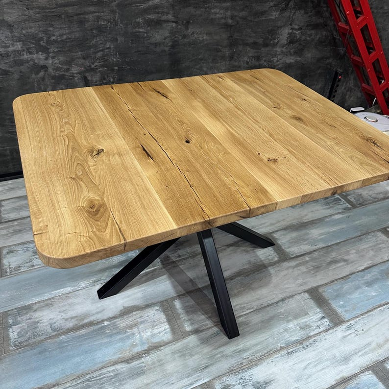 A rectangular wooden dining table with a natural oak top and a black metal X-frame base, shown in an industrial warehouse setting with gray concrete walls and distressed gray-blue flooring.
