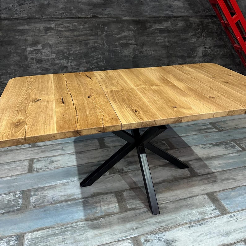 A rectangular wooden dining table with a light brown oak top and a black metal X-base pedestal, shown in an industrial warehouse setting with a dark gray concrete wall and distressed gray wood floor.