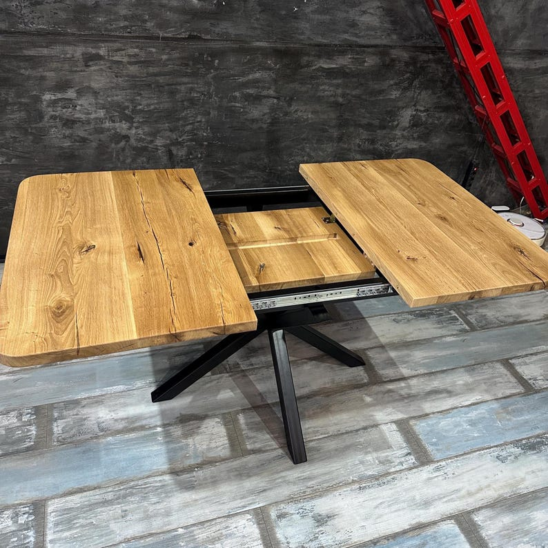 A rustic wooden extendable table with a black metal frame, shown with both side panels partially extended to reveal the interior mechanism. The tabletop has a natural light brown wood finish with visible grain and knots, and the base is a black metal cross-style leg.