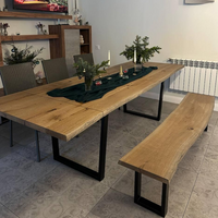 A rustic wooden dining table with a live-edge oak top and black metal legs, accompanied by a matching wooden bench and woven dining chairs, set in a dining room.