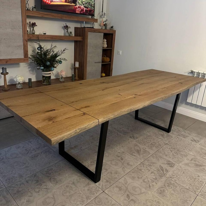 A rustic rectangular wooden dining table with a live-edge slab top and black metal frame legs, shown in a home interior setting with tiled flooring and wooden cabinetry in the background.