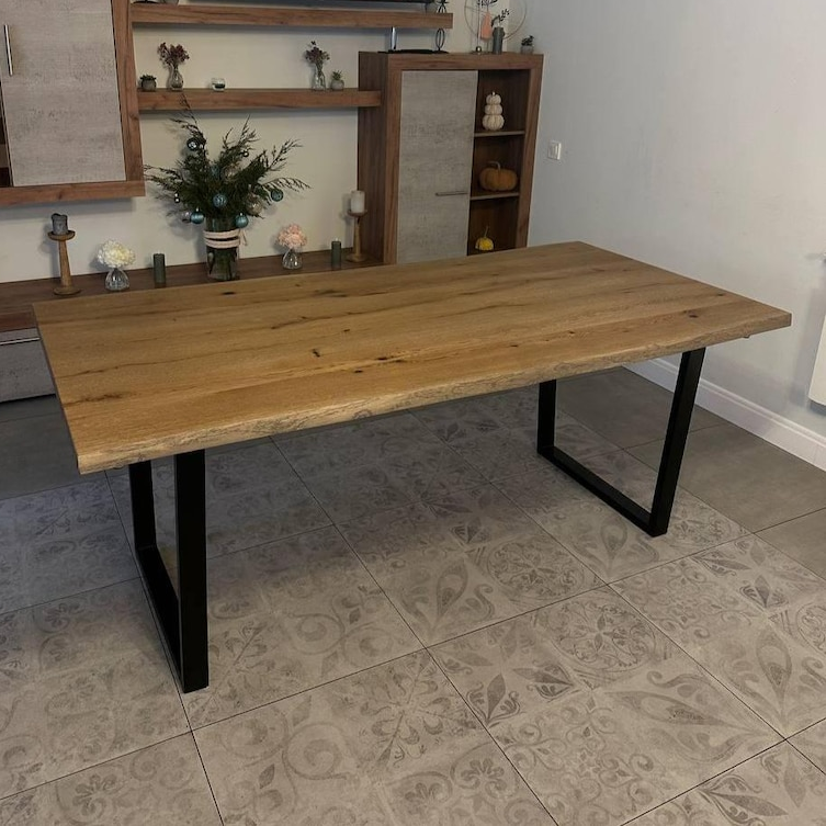 A rustic live-edge wooden dining table with a natural oak top and black metal legs, shown in a home interior setting with tiled flooring and wooden shelving in the background.