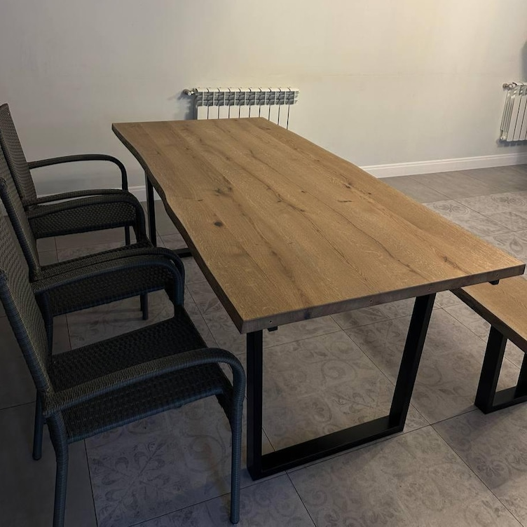 A rectangular wooden dining table with a natural oak top and black metal legs, shown alongside black woven armchairs in a room with tiled floors and a white wall.
