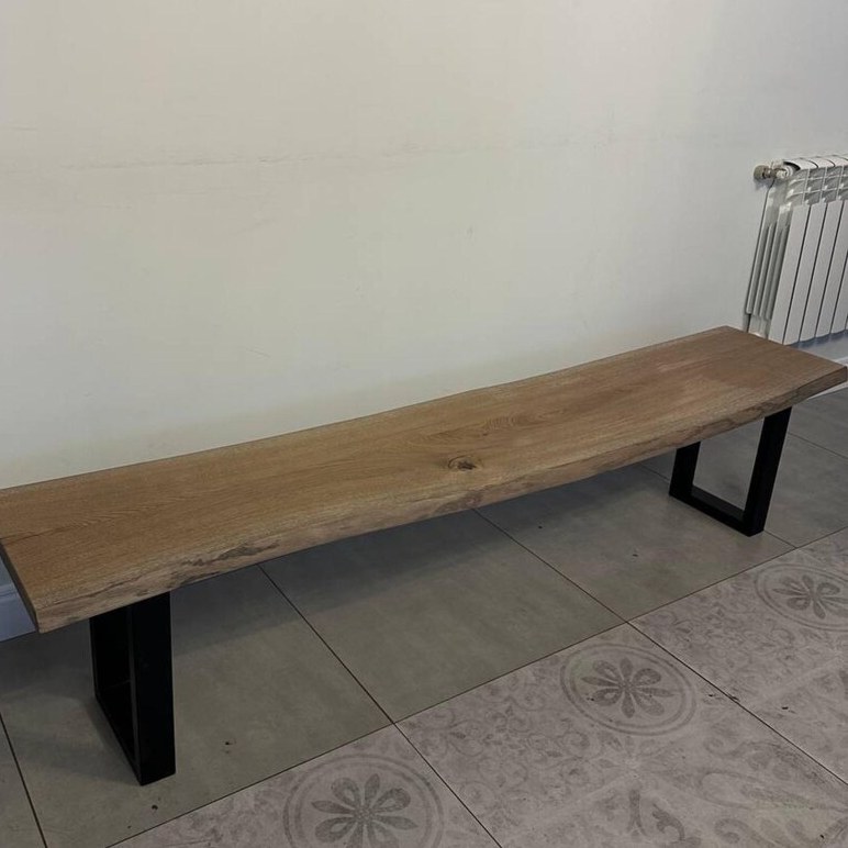 A live-edge wooden bench with a natural oak tabletop and black metal legs, shown in a room with tiled flooring and a white wall.
