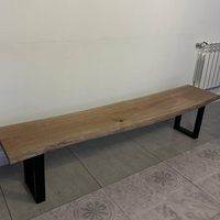 A live-edge wooden bench with a natural oak tabletop and black metal legs, shown in a room with tiled flooring and a white wall.