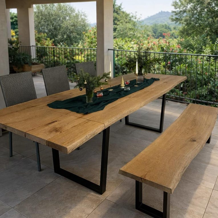 A large rectangular outdoor dining table with a natural oak wood top and black metal legs, accompanied by a matching wood bench and woven chairs, set on a tiled patio with a lush garden background.