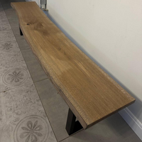 A rustic live-edge wooden bench with a natural oak top and black metal T-leg base, photographed in an indoor setting with tiled flooring and a white wall.